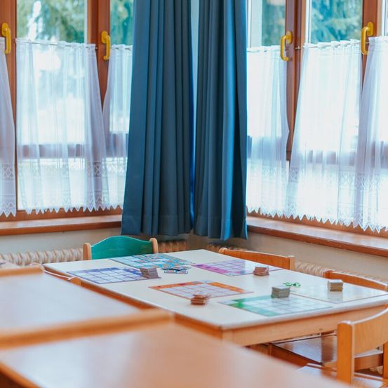 Ein leerer Klassenraum mit einem Holztisch und Stühlen, ausgestattet mit blauen Vorhängen, weißen Vorhängen und Fenstern mit Blick auf Bäume draußen.