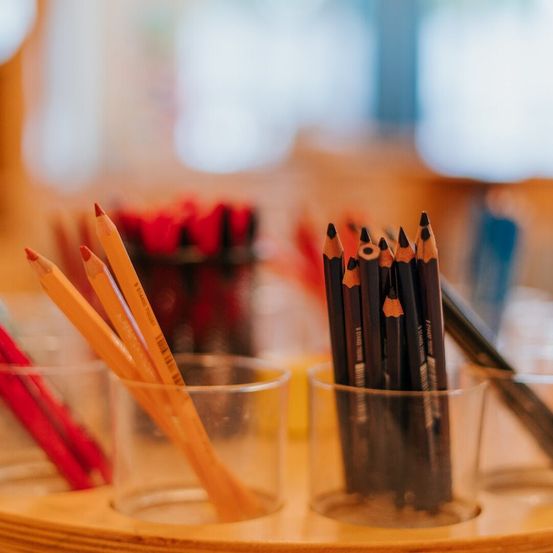 Zwei durchsichtige Glasbehälter halten Bleistifte. Ein Behälter hat rote Bleistifte und der andere hat schwarze Bleistifte. Unscharfe Objekte und ein Fenster sind im Hintergrund.