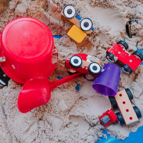 Verschiedene bunte Spielzeuge, darunter ein roter Eimer und ein lila Becher, liegen verstreut in einem Sandkasten mit Sand. Es gibt auch kleine hölzerne Spielzeugfahrzeuge mit Rädern und Augen.