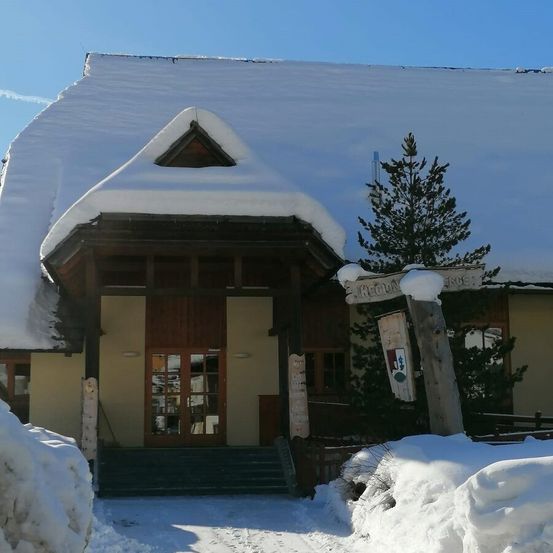 Ein schneebedecktes Gebäude mit einem dreieckigen Dacheingang, Glastüren und einem Schornstein. Schneehaufen umgeben das Gebäude und eine Tanne steht in der Nähe.