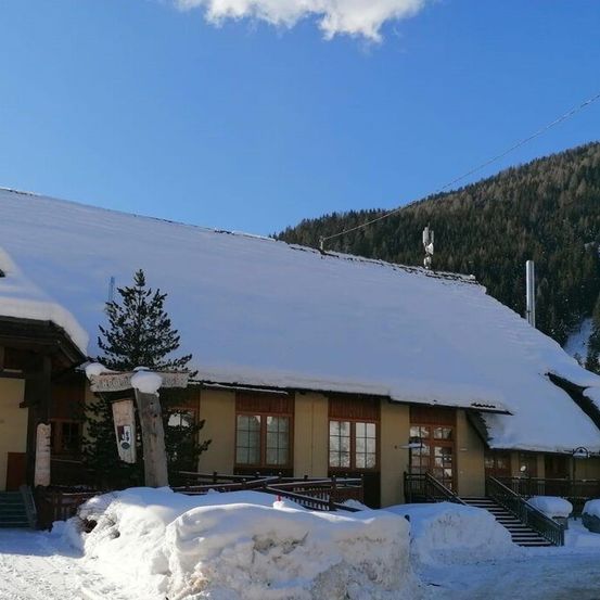Ein schneebedecktes Gebäude mit einem steilen Dach und mehreren Fenstern steht in einer verschneiten Landschaft mit einem Berg im Hintergrund.