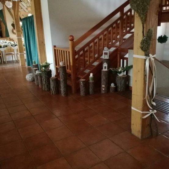 Ein Innenbereich mit braun gefliestem Boden, einer Holztreppe und dekorativen Baumstämmen mit Pflanzen und Kerzen darauf. Es gibt einen blauen Vorhang und einen kleinen Tisch in der Ferne.