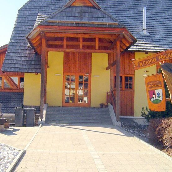 Ein Holzgebäude mit einem Schieferdach und gelben Wänden verfügt über einen hölzernen Vorbau. Links stehen zwei Mülleimer. Ein orangefarbener Banner mit Text hängt über dem Eingang.