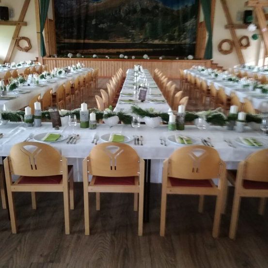 Ein großer Speisebereich mit mehreren Tischen und Stühlen, geschmückt mit weißen Tischdecken, Kerzen und Grünpflanzen. Der Raum verfügt über Holzböden und Wände mit einem großen Wandbild.