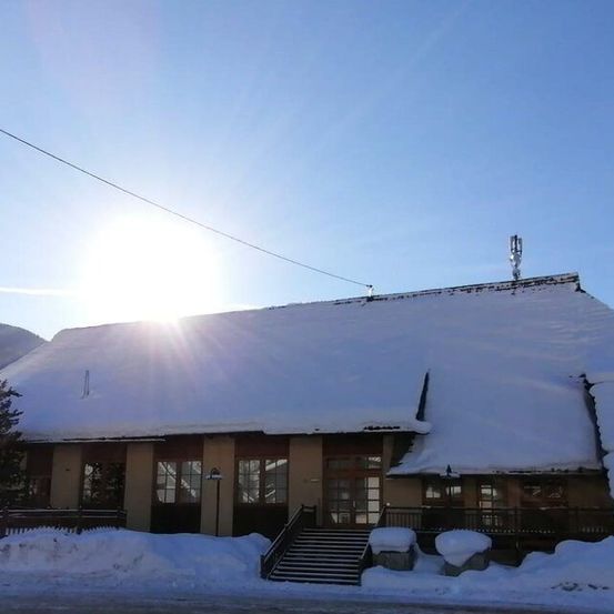 Ein Gebäude mit schneebedecktem Dach steht in einer verschneiten Landschaft unter einem strahlenden Sonnenschein.