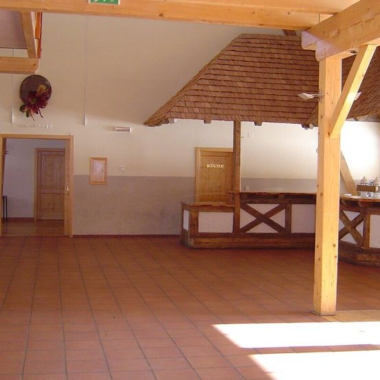 Ein leerer Raum mit Holzsäulen, einem gefliesten Boden und einer hölzernen Theke mit der Aufschrift 'Köche'. Eine Tür führt zu einem offenen Flur. Ein Korb mit Blumen hängt an der Wand.