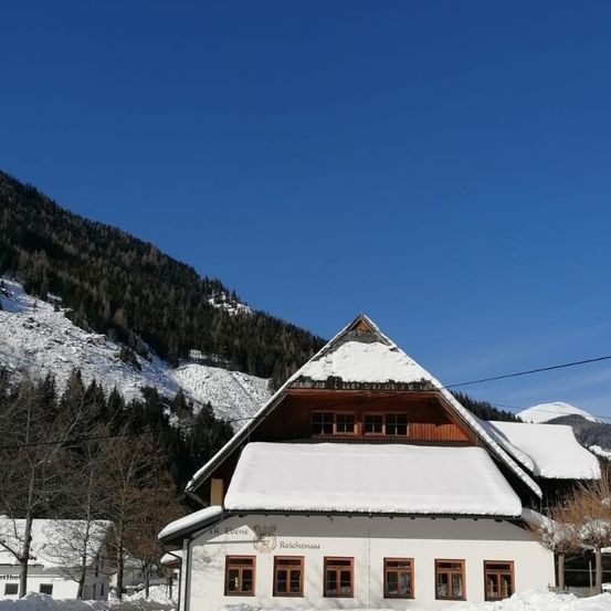 Ein schneebedecktes Haus mit einem dreieckigen Dach und mehreren Fenstern befindet sich in einem verschneiten Gebirgsgebiet. Das Haus ist schneebedeckt und umsäumt von Bäumen. In der Ferne erstreckt sich ein schneebedeckter Bergkamm.