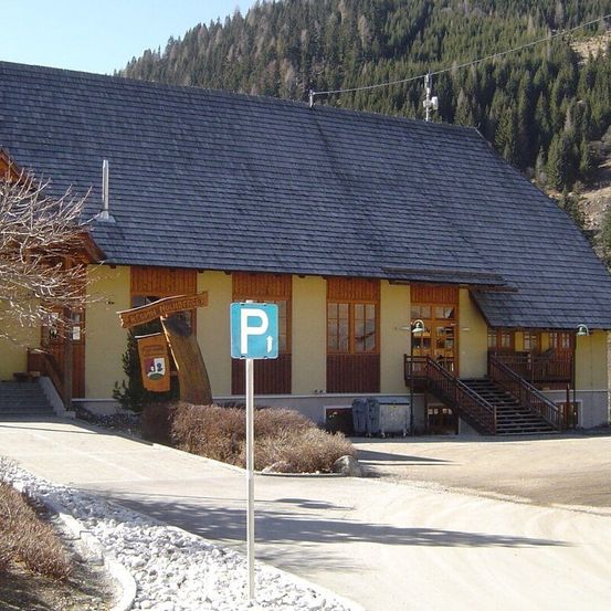 Ein Gebäude mit einem Schieferdach, gelben Wänden und Holzverkleidungen steht in einem bergigen Gebiet. Ein Schild mit dem Buchstaben P befindet sich auf der Einfahrt. Bäume umgeben das Gebäude und es gibt eine Treppe, die zum Eingang führt.