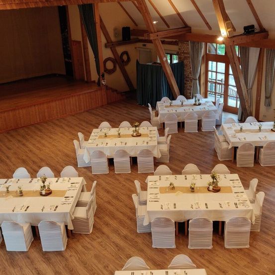 Ein Indoor-Essbereich mit Holzböden, eingerichtet mit Tischen und Stühlen, mit einer Bühne an einem Ende und Holzbalken darüber. Jeder Tisch ist mit kleinen Pflanzen und Besteck geschmückt.