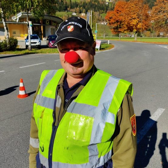 Ein Mann mit roter Clownnase und reflektierenden Weste und Hut steht auf der Straße, nahe einem orangefarbenen Verkehrskegel.