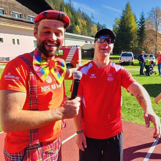 Zwei Männer in roten T-Shirts und Sonnenbrillen stehen auf einer Laufbahn, einer hält ein Mikrofon. Einer trägt einen Kilt und eine bunte Schleife. Im Hintergrund steht ein Gebäude und es sind Autos geparkt.