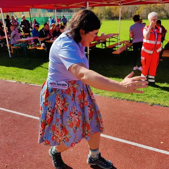 Eine Frau in einem Blumenrock mit einem Namensschild, das Rote Nasen Clowndoctors steht, steht auf einer Laufbahn und gestikuliert mit ihren Händen. Hinter ihr bedeckt eine Frau in einer reflektierenden Weste ihren Mund. Im Hintergrund sitzen Menschen unter einem Zelt, und einige stehen in der Nähe von Tischen.