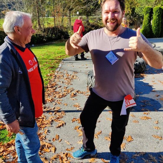 Zwei Männer in einer Außenumgebung, einer lächelt und gibt Daumen hoch, der andere mit einem roten Shirt und blauer Jacke.