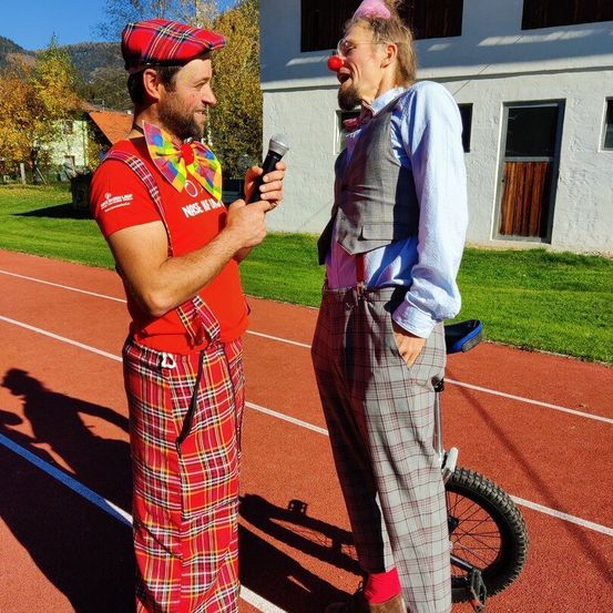 Zwei Männer stehen auf einer Laufbahn. Ein Mann hält ein Mikrofon und interviewt den anderen Mann, der auf einem Einrad steht und eine rote Clownnase trägt.