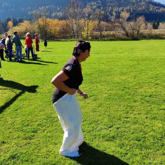 Eine Frau läuft auf einem Grasfeld, umgeben von mehreren Menschen, die in einer Reihe im Hintergrund stehen. Die Bäume sind in voller Farbe, und die Berge sind in der Ferne.