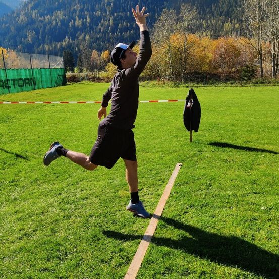 Ein Mann auf einem Rasenfeld wirft einen Ball, während er auf einer Holzplanke steht. Er trägt einen Hut, ein Langarmshirt, Shorts und Sneaker. Hinter ihm sind ein Zaun und Bäume zu sehen.