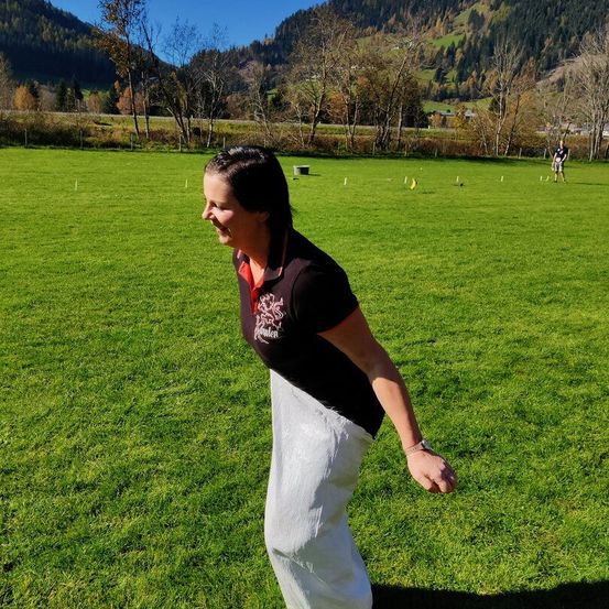 Eine Frau in einem schwarzen und roten Shirt und weißen Hosen steht auf einem Feld mit Bergen im Hintergrund.