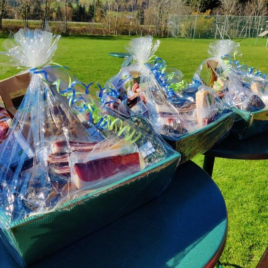 Mehrere verpackte Geschenkkörbe stehen auf einem blauen Tisch im Freien. Jeder Korb enthält verschiedene Artikel, darunter Fleisch, Käse und andere Lebensmittel. Die Körbe sind in Plastik mit Schleifen verpackt.