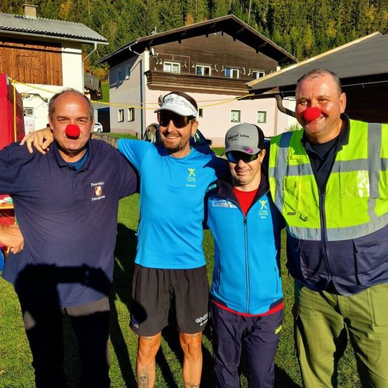 Vier Menschen in Outdoor-Kleidung stehen auf Gras. Zwei Männer tragen rote Clownsnasen. Gebäude und Bäume sind im Hintergrund.
