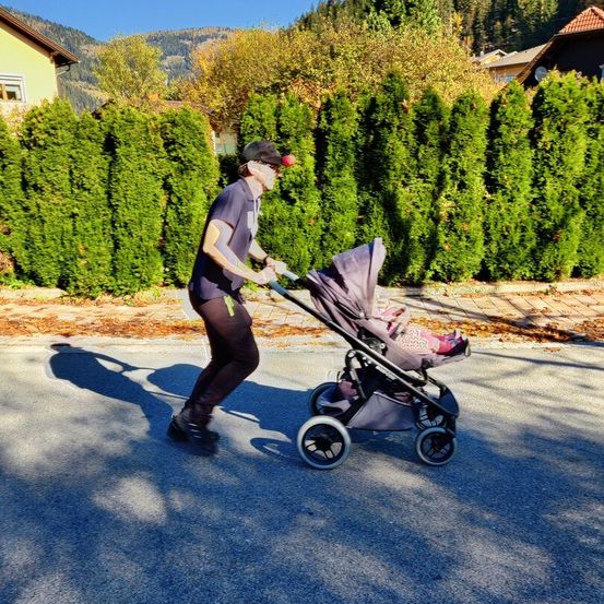 Ein Mann schiebt einen Kinderwagen die Straße hinunter, mit einem Baby darin. Der Weg ist gesäumt von hohen, grünen Hecken und Bergen im Hintergrund.