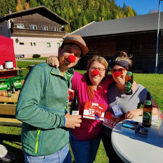 Drei Menschen mit roten Nasen posieren für ein Foto in einem Park mit einem Gebäude und Bäumen im Hintergrund. Einer hält eine Flasche Bier, und ein Tisch mit Getränken ist in der Nähe.