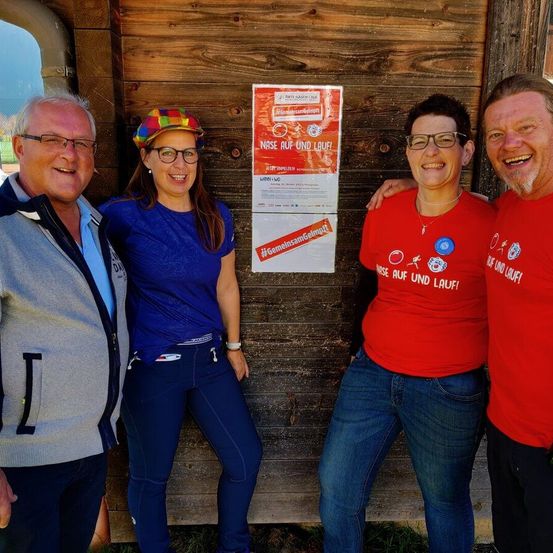Vier Personen, die lächeln und für ein Foto posieren, stehen vor einer Holzwand mit einem roten Plakat mit Text und Symbolen.