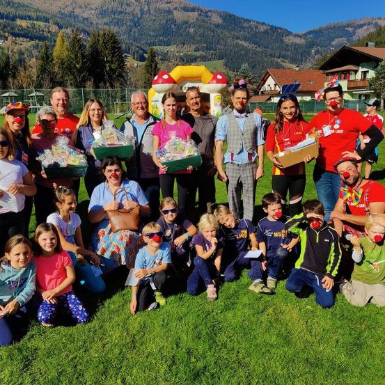 Eine Gruppe von Erwachsenen und Kindern posiert auf einem Rasenfeld vor einem Bergpanorama. Einige Erwachsene halten Boxen und einige Kinder tragen rote Clownsnasen.