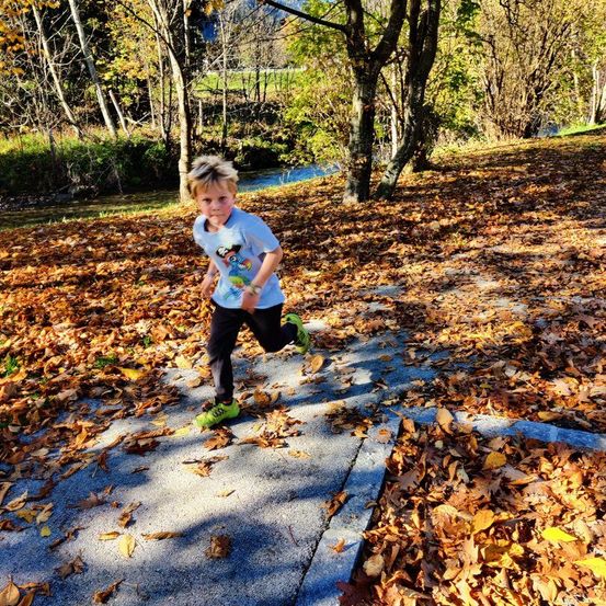 Ein junger Junge läuft auf einem Pfad mit gefallenen Blättern. Er trägt ein weißes Shirt und grüne Turnschuhe. Der Pfad ist von Bäumen und Gras umgeben.
