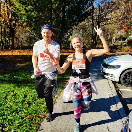 Ein Mann und eine Frau, beide lächelnd, laufen Hand in Hand einen Bürgersteig hinunter. Der Mann trägt eine blaue Kappe und ein weißes T-Shirt, während die Frau ein schwarzes Tanktop und bunte Leggings trägt. Hinter ihnen steht ein weißes Auto, Bäume und ein Rasenbereich.