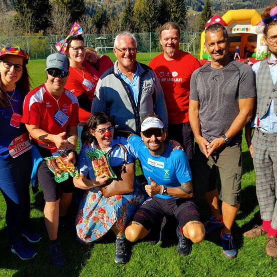 Eine Gruppe von Menschen in bunter und festlicher Kleidung versammelt sich auf einem Rasenfeld, möglicherweise für eine Wohltätigkeitsveranstaltung. Sie lächeln und posieren für ein Foto. Einige halten Lebensmittel in den Händen, während andere eine Brille und einen Hut tragen. Im Hintergrund befindet sich ein Hüpfburg und ein Zaun.