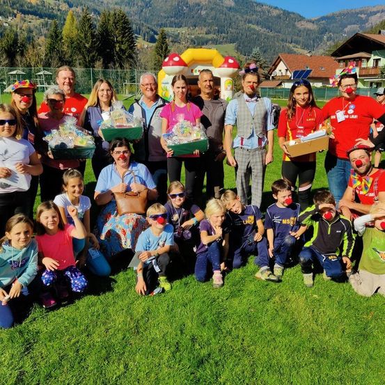 Eine Gruppe von Erwachsenen und Kindern, einige mit Clown-Make-up, posieren auf einem Rasenfeld mit Bergen im Hintergrund. Einige halten Boxen.