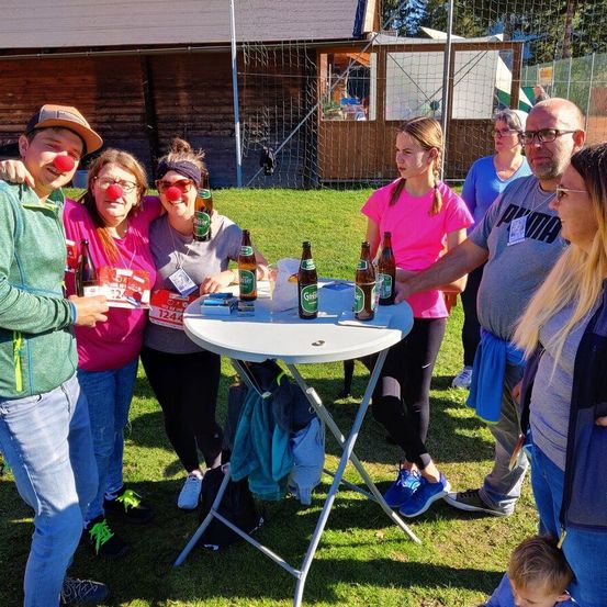 Eine Gruppe von Menschen steht um einen Tisch mit Flaschen darauf. Sie tragen Freizeitkleidung. Ein Mann trägt eine Brille und hält eine Flasche. Die Frau davor trägt ein rosa Shirt.