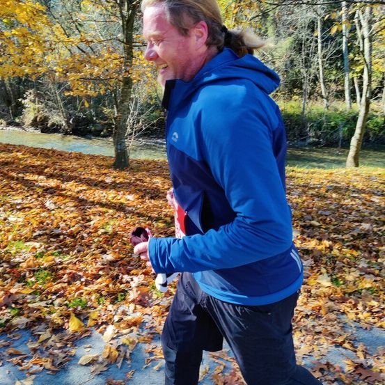 Ein Mann läuft im Freien in einem blauen Jacken und schwarzen Hosen. Er lächelt und hält einen roten Gegenstand. Der Boden ist mit gefallenen Blättern bedeckt. Bäume und ein Fluss sind im Hintergrund.