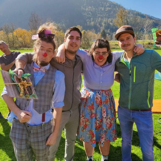 Eine Gruppe von vier Erwachsenen, die Clown-Make-up und Kostüme tragen, posiert für ein Foto auf einer Grasfläche vor dem Hintergrund von Bäumen und Bergen.