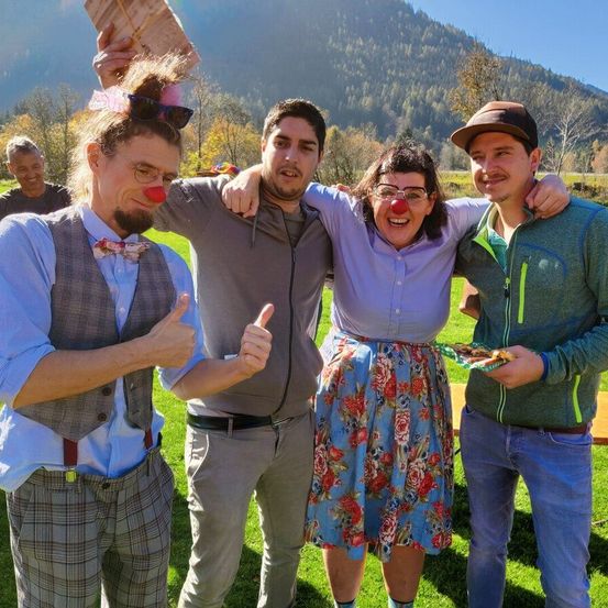 Vier Personen posieren für ein Foto auf einer Wiese mit Bergen im Hintergrund. Einer trägt eine Clownnase und einen Fliegenbinder, ein anderer eine Papierkrone und ein Dritter hält Essen.