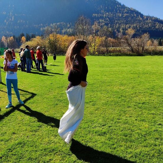 Eine Frau mit langen Haaren läuft auf einem Grasfeld mit einem weißen Tuch. Ein Mädchen steht in der Nähe. Leute stehen in einer Reihe. Ein Berg mit Bäumen und Büschen ist in der Ferne.