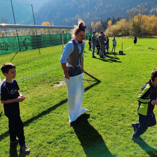Ein Mann in einem bunten Kostüm steht auf einem Feld. Kinder beobachten, einer läuft. Ein Fußballnetz im Hintergrund, eine Gruppe von Menschen beobachtet.