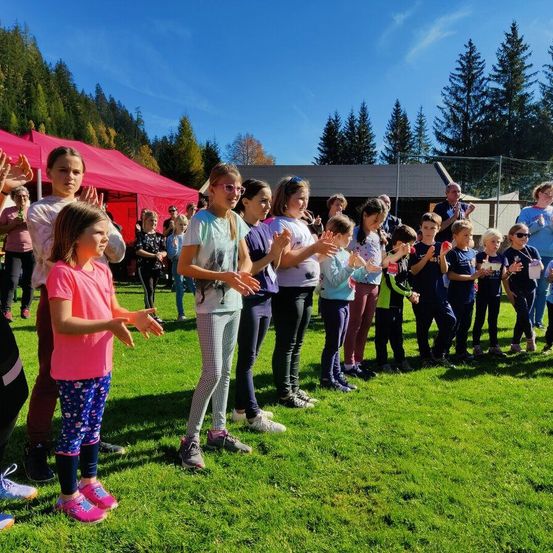 Kinder in Freizeitkleidung stehen auf einem grünen Feld, möglicherweise führen sie einen Tanz oder eine Cheerleading-Aufführung durch. Sie stehen vor der Kamera, dahinter befindet sich ein Zelt und mehrere Personen. Im Hintergrund sind Bäume und ein Gebäude zu sehen.