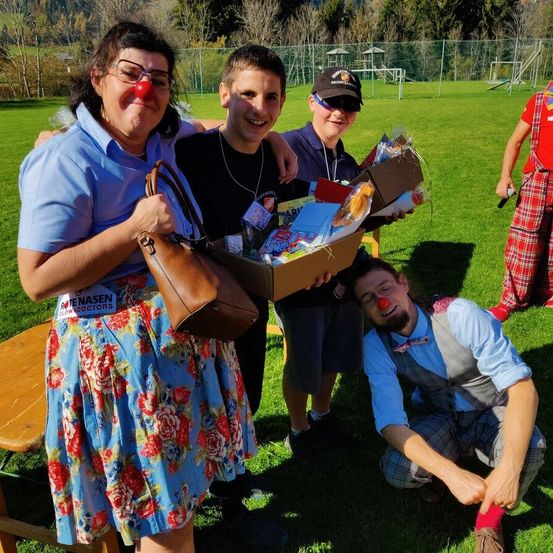 Vier Personen lächeln auf einer Wiese. Ein Mann hält eine Schachtel mit verschiedenen Gegenständen, und ein anderer hockt mit einer roten Nase. Eine Frau mit Brille und einem Blumenrock hält eine braune Tasche. Eine Person in einem roten Karorock steht hinter ihnen.