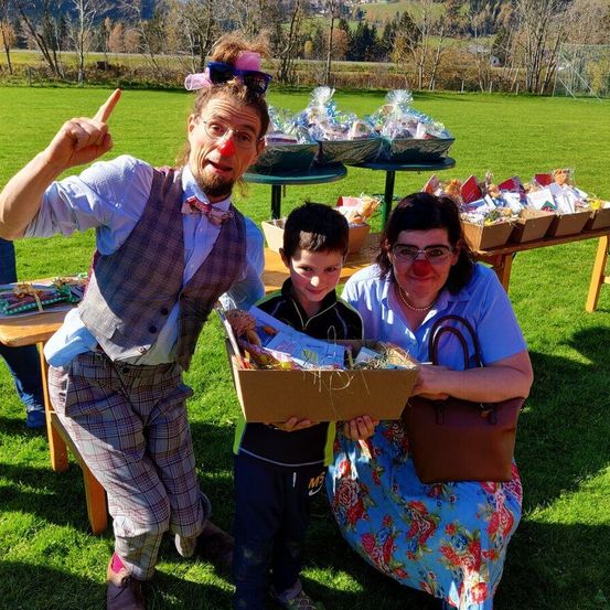 Drei Personen posieren für ein Foto. Ein Mann in Clown-Kleidung und eine Frau in einem Blumenkleid halten eine Geschenkbox, während ein Junge die Box in den Händen hält. Sie stehen auf einer Grasfläche mit Tischen voller Geschenke.