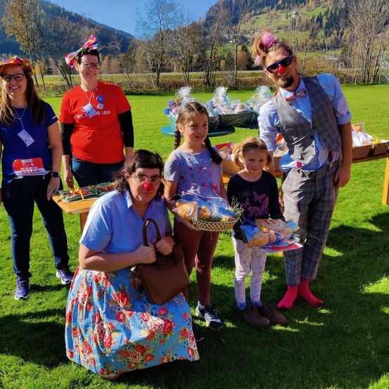 Eine Gruppe von Menschen in Kostümen steht auf einer Wiese. Zwei Mädchen halten Körbe mit Essen. Ein Mann mit Fliege und eine Frau mit roter Nase sind ebenfalls anwesend. Hinter ihnen stehen Tische mit verpackten Geschenken. Im Hintergrund sind Berge und Bäume zu sehen.