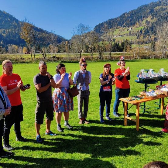 Eine Gruppe von Menschen steht auf einer Wiese, einige tragen Hüte und Brillen. Ein Tisch mit verschiedenen Gegenständen ist in der Nähe. Im Hintergrund befinden sich Berge und Bäume.