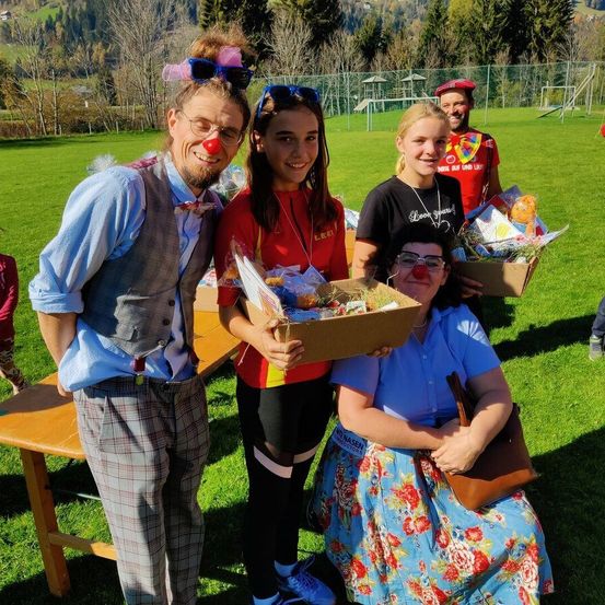 Eine Gruppe von Menschen in Clownkostümen und mit Geschenkboxen posiert für ein Foto auf einer Wiese mit einer Holzbank und Bäumen im Hintergrund.