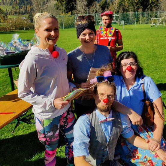 Eine Gruppe von Menschen posiert für ein Foto auf einem Rasenfeld. Eine Frau mit bunten Leggings hält ein Buch. Eine andere Frau mit roter Clownsnase sitzt auf dem Schoß eines Mannes. Dahinter steht ein Holztisch mit einem Korb und einer Person in Clownskostüm. In der Ferne gibt es Bäume und einen Spielplatz.