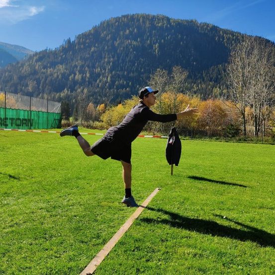 Ein Mann in Sportbekleidung übt das Werfen eines Gegenstands auf einem Rasenfeld, mit Bergen und Bäumen im Hintergrund.