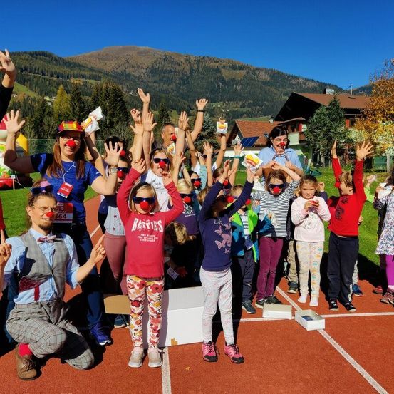 Eine Gruppe von Menschen mit Clownmasken und Sonnenbrillen posiert für ein Foto auf einer Laufbahn mit Bergen im Hintergrund.