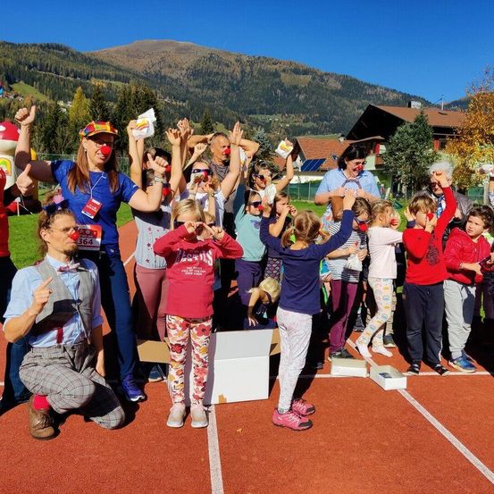 Eine Gruppe von Erwachsenen und Kindern in Kostümen steht auf einer Laufbahn und trägt rote Nasen, Brillen und hält einige Snacks. Sie scheinen für ein Foto zu posieren.