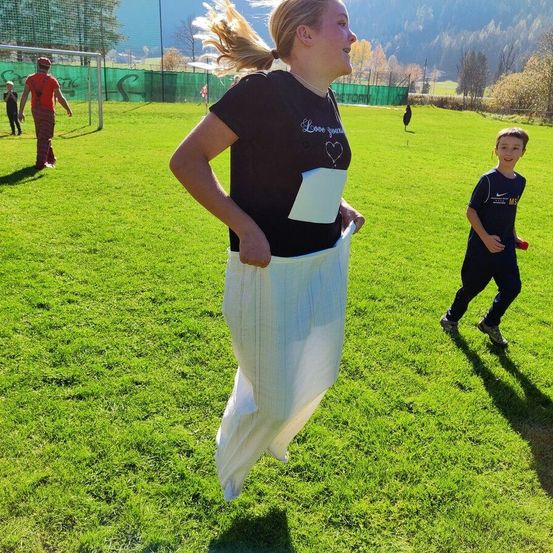 Eine Frau in einem schwarzen T-Shirt und weißen Hosen läuft auf einem Grasfeld, während ein Junge in Blau in der Nähe joggt. Im Hintergrund steht ein Mann in der Nähe eines Fußballtors.