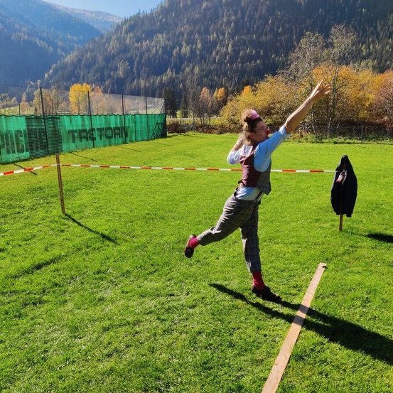 Eine Person wirft einen Speer im Freien an einem sonnigen Tag. Der Bereich ist mit einem grünen Netz und roten Seilen eingezäunt. Bäume und Berge sind im Hintergrund.