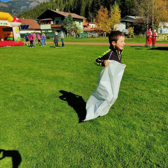 Ein Junge rennt auf einem Rasenfeld und hält ein weißes Tuch. Hinter ihm ist ein aufblasbarer Pilz zu sehen. Im Hintergrund sind Häuser und Bäume zu erkennen.